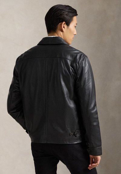 Homme vu de dos portant une veste en cuir noire et un pantalon noir sur un fond clair uni.