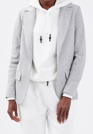 Personne portant un blazer gris clair par-dessus un sweat à capuche blanc avec des cordons noirs et un pantalon blanc, debout devant un fond uni.