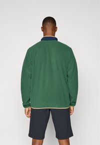 Veste en polaire verte avec un col en marine contrastant et des finitions beiges, présentant une coupe décontractée et des manches longues. Portée avec un short noir.