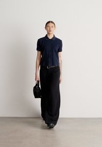 Tumši zils adīts īss piedurkņu polovers, melnas platu kāju bikses, rokās tur mazu melnu somu, stāvot pret vienkāršu balto fonu.