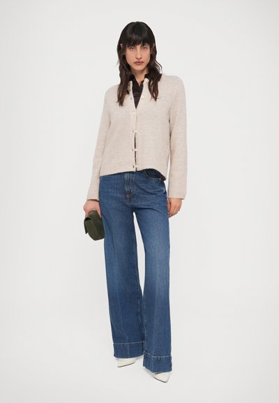 Femeie purtând un cardigan bej cu nasturi, cămașă neagră, blugi albaștri largi, pantofi albi cu vârf ascuțit, ținând o poșetă mică verde.