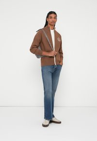 Brun zip-up genser med høy krage og hvit tekst, kombinert med blå jeans og kremfargede sneakers, holder en grå bag over skulderen.
