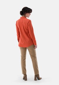 Rust-orange long-sleeve blouse with a collar, paired with beige tailored pants. Details include buttons on the sleeves and a smooth texture.