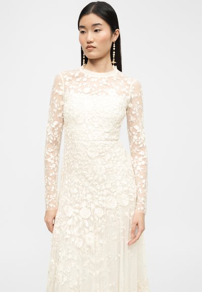 Νεαρή γυναίκα με μακρυμάνικο λευκό φόρεμα από δαντέλα με λουλούδια και μακριά σκουλαρίκια με πέρλες και αστεροειδές σχέδιο, στέκεται μπροστά σε απλό φόντο.