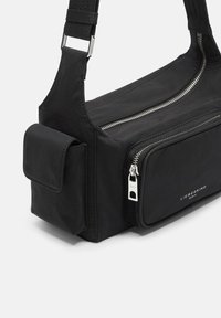 Sac à épaule en tissu noir avec un compartiment principal zippé, une poche frontale et des détails en métal argenté. Équipé d'une bandoulière large et réglable.