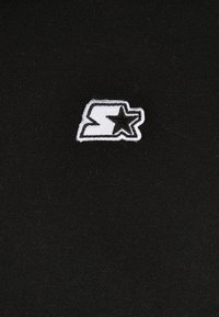 Tela negra con un logo bordado. El diseño incluye una letra "S" estilizada con una estrella, en hilo blanco, sobre una textura suave.