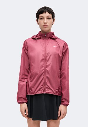 Henkilö, jolla on päällään viininpunainen Nike-huppari vetoketjulla ja musta hame, seisoo kasvot eteenpäin yksiväristä taustaa vasten.