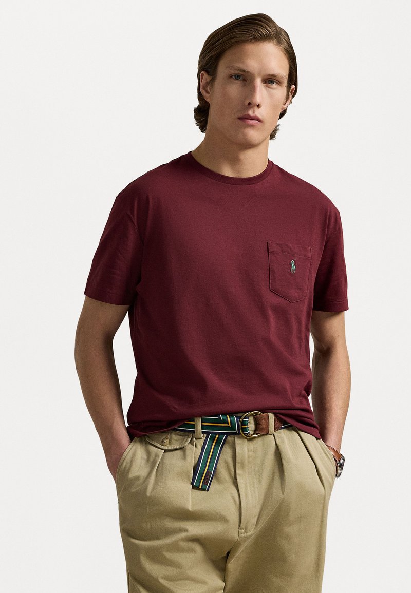 Burgundy bomull t-shirt med rund hals, korta ärmar och en bröstficka med en liten logotyp. Kombinerad med khakifärgade byxor och ett randigt bälte.