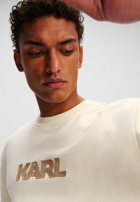 Lys beige sweatshirt lavet af blødt stof, med et fremtrædende brunt broderet "KARL" logo på brystet og en ribbet rund hals.
