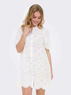 Jeune femme portant une robe blanche à œillets boutonnée, manches courtes, souriante et regardant vers le bas, main près du cou.