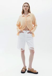 Camicia arancione chiaro a maniche lunghe con una sola tasca, pantaloni corti bianchi con orlo sfrangiato e sandali marroni con suola nera e decorazioni dorate.