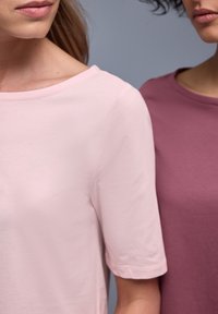 Deux femmes se tiennent côte à côte, portant des t-shirts unis à col rond, l'un rose clair et l'autre mauve, sur un fond neutre.