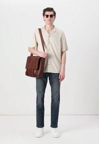 Camisa de botones de manga corta de color claro, jeans de mezclilla oscura, zapatillas blancas y un bolso de mensajero de cuero marrón. Los accesorios incluyen gafas de sol oscuras.