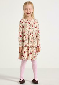 Uśmiechnięta młoda dziewczynka stoi, ubrana w beżową kwiecistą sukienkę, różowe rajstopy w wzory i ciemnobrązowe plecione buty na jednolitym tle.