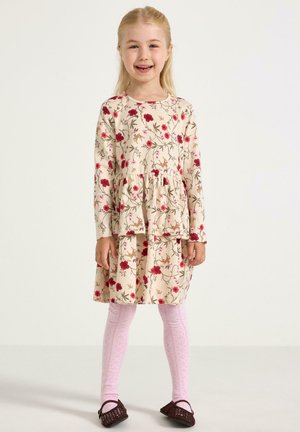 Uśmiechnięta młoda dziewczynka stoi, ubrana w beżową kwiecistą sukienkę, różowe rajstopy w wzory i ciemnobrązowe plecione buty na jednolitym tle.