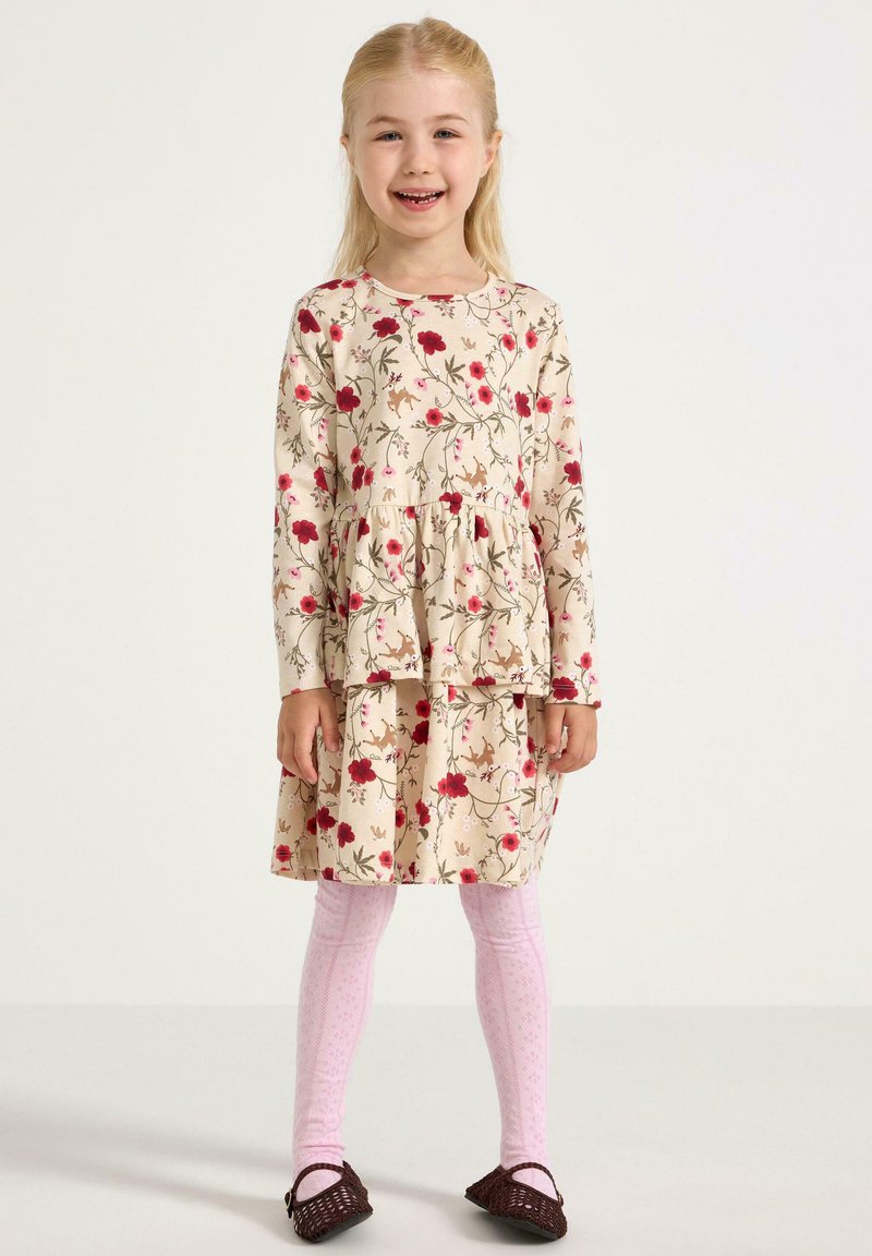 Uśmiechnięta młoda dziewczynka stoi, ubrana w beżową kwiecistą sukienkę, różowe rajstopy w wzory i ciemnobrązowe plecione buty na jednolitym tle.