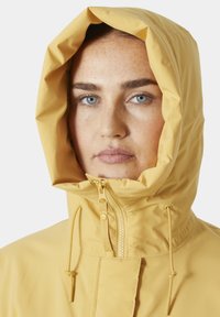 Chaqueta impermeable amarilla con capucha ajustable, cremallera frontal y textura suave, presenta un diseño minimalista y sin patrones visibles.