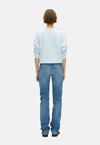 Personne avec les cheveux bruns courts portant un sweat-shirt bleu clair et un jean bleu droit, se tenant de dos sur un fond uni.