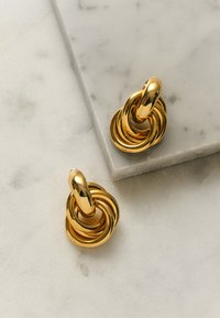 Goldfarbene Ohrringe mit glattem, glänzendem Finish und ineinander verschlungenen Schlaufen. Auf einer hellen Marmoroberfläche platziert.