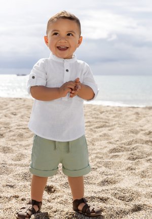 Dítě oblečené v bílé lněné košili s vyhrnutými rukávy, zelených krátkých kalhotách a hnědých sandálech, stojící na písčité pláži s vodou v pozadí.