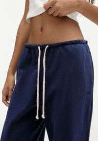 Kvinde iført marineblå sweatpants med hvide snore og en cropped hvid top, hånd justerer toppen nær taljen.