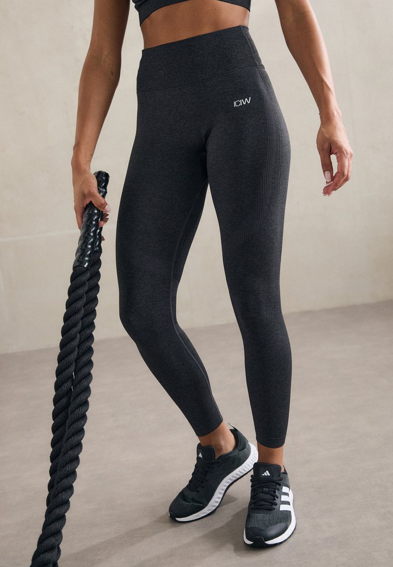 Høyt liv, mørkegrå leggings med teksturert mønster og logo, kombinert med svarte treningssko, holder en tykk svart tau.