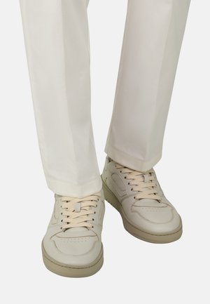 Piedi con indosso sneakers beige chiaro con lacci abbinate a pantaloni color crema su sfondo bianco.
