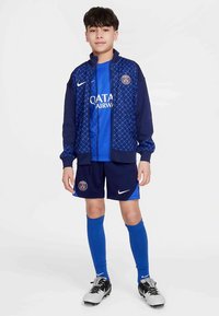 Blåt Nike sportssæt med en mønstret jakke og matchende skjorte, shorts, sokker og fodboldstøvler, med et logo på brystet og shorts.