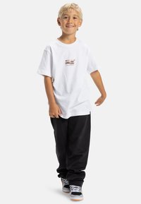 Niño sonriente con cabello rubio que lleva una camiseta blanca con un gráfico de una cámara vintage, pantalones negros y zapatillas en blanco y negro.