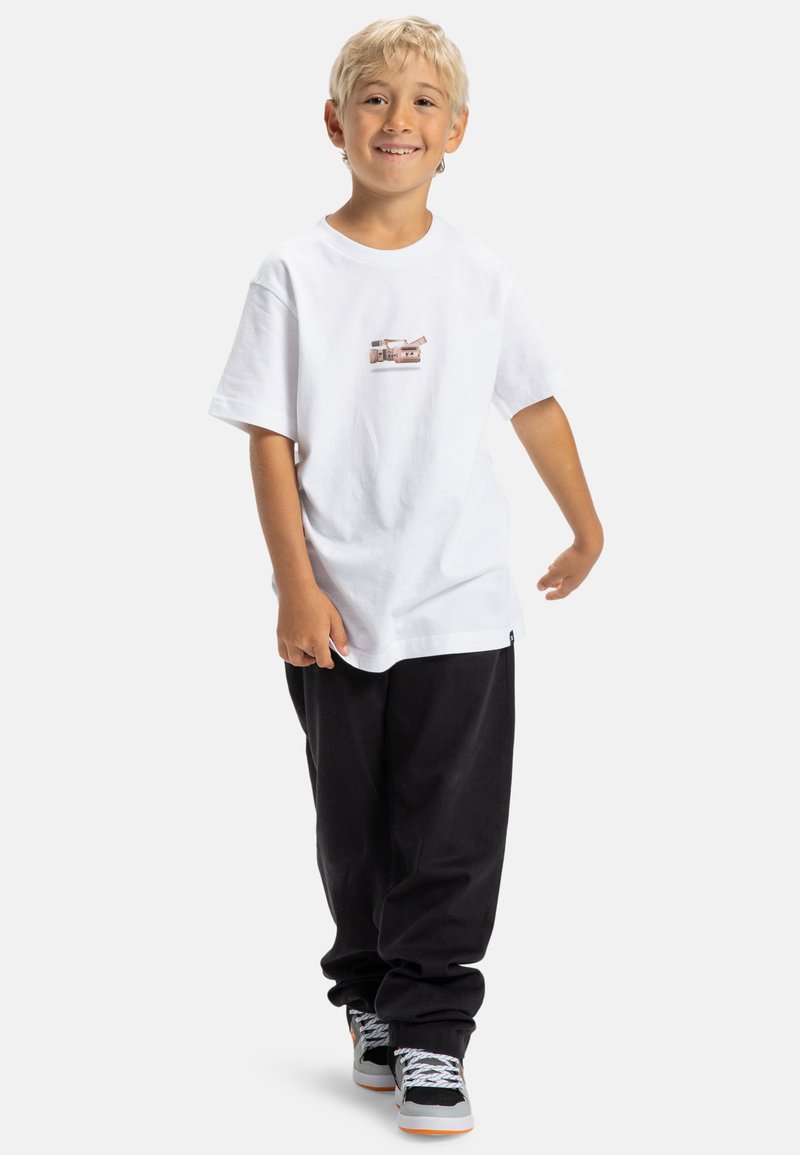 Niño sonriente con cabello rubio que lleva una camiseta blanca con un gráfico de una cámara vintage, pantalones negros y zapatillas en blanco y negro.