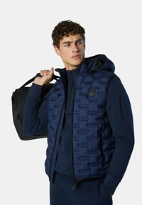 Marineblaue Steppweste mit Kapuze, gestepptem Muster, Reißverschluss vorne und Seitentaschen, getragen über einem passenden, figurbetonten Oberteil, begleitet von einer schwarzen Tasche.