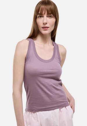 Femme aux cheveux bruns raides portant un haut mauve sans manches et un pantalon rose clair, debout avec une main dans la poche sur un fond uni.