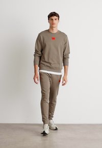 Felpa in cotone grigia con patch logo rosso, abbinata a pantaloni jogger grigi coordinati. Sneakers grigie completano l'outfit. Texture liscia.