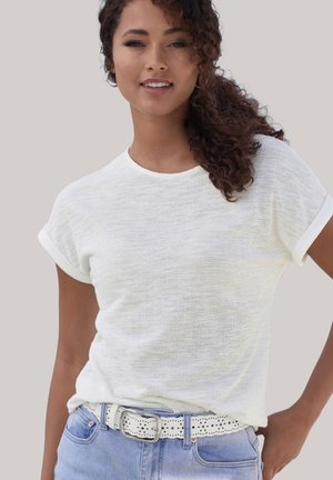 Frau mit lockigem Haar, die ein weißes, kurzärmliges, strukturiertes Oberteil und hellblaue Jeans mit einem weißen, gelochten Gürtel vor neutralem Hintergrund trägt.