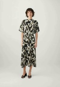 Žena stoji, odjevena u midi haljinu s crno-bijelim cvjetnim uzorkom, širokim rukavima i crnim sandalama na blok petu, protiv jednostavne svijetle pozadine.