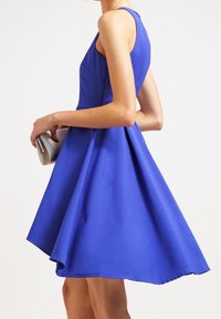 Bezruké šaty do kolen v jasně modré barvě s rozšířenou sukní a hladkou látkou. Modelka drží malou stříbrnou kabelku.