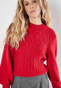 Czerwony sweter z dzianiny z teksturowaną górą z diamentowymi wzorami, prążkowanym kołnierzem i dolną częścią w warkocze. Stylizowany z szarymi spodniami.