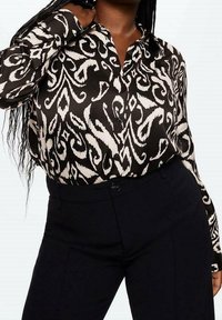 Blusa a fantasia bianco e nero con colletto, maniche lunghe e dettagli di bottoni discreti, abbinata a pantaloni scuri.