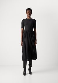 Donna in un abito midi plissettato nero con maniche a lunghezza gomito e stivali neri con tacco, su uno sfondo bianco semplice.