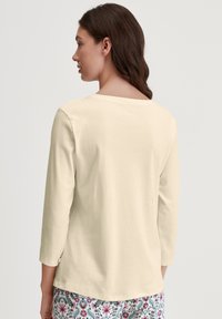 Crème-kleurige blouse met lange mouwen en een ronde halslijn, gladde textuur en schoudernaden. Gekocht met bedspijkers met een patroon van bloemontwerpen.