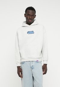Sudadera con capucha gris y logo gráfico azul en el pecho, bolsillo frontal y puños de canalé. Usada con pantalones de mezclilla azul claro.
