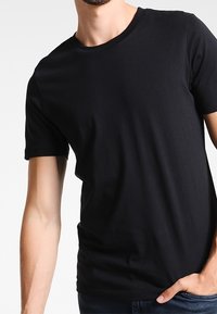 Muž oblečený v jednoduchém černém tričku s krátkým rukávem a kulatým výstřihem, s jednou rukou v kapse, na bílém pozadí.