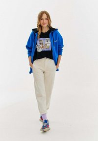 Niebieska bluza rozpinana z kapturem, czarna koszulka z graficznym nadrukiem w kolorowy wzór, kremowe spodnie i wielokolorowe sneakersy. Gładkie tekstury w całym zestawie.