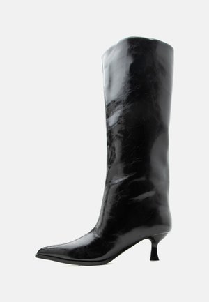 Botte en cuir noire brillante, haute jusqu'au genou, avec bout pointu et talon bas courbé sur un fond clair uni.