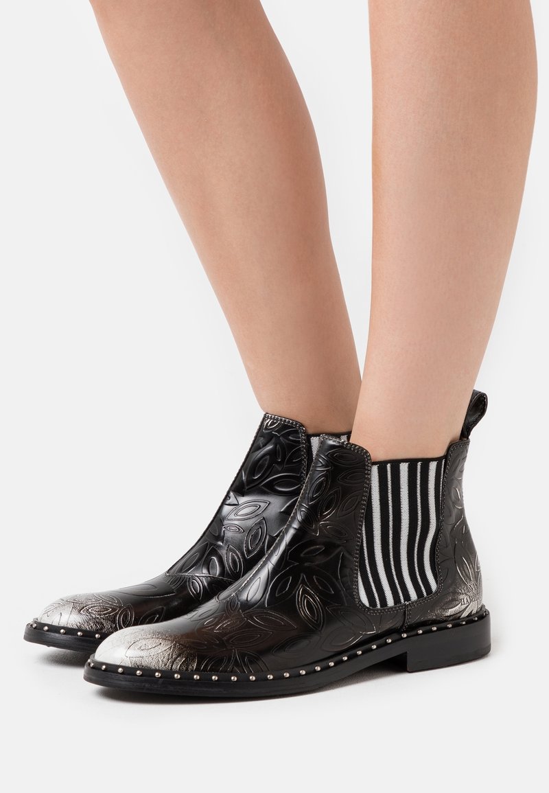 Bottines noires avec motif de feuilles embossé, embout argenté, panneaux latéraux élastiques à rayures noires et blanches, et clous décoratifs argentés sur la semelle.