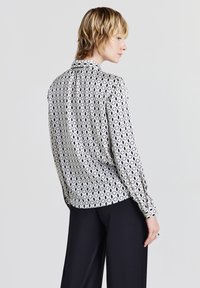 Langärmlige Bluse mit Kragen im schwarz-weißen Muster mit ovalen Formen. Leichter Stoff, glatte Textur, sichtbare Knopfmanschetten und taillierter Schnitt.