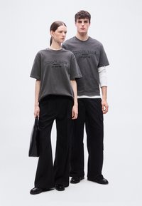 Δύο μοντέλα στέκονται δίπλα δίπλα φορώντας παρόμοια ξεθωριασμένα σκούρα γκρι μπλουζάκια Tommy Jeans και μαύρο παντελόνι, το ένα κρατά μια μαύρη τσάντα.