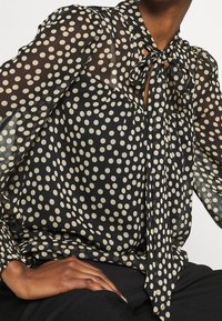 Blusa nera trasparente con grandi pois crema, maniche lunghe, dettaglio al collo con fiocco e vestibilit�à ampia. Il tessuto sembra morbido e leggero.
