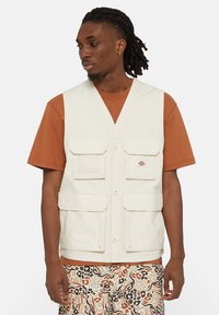 Gilet utilitario beige con più tasche, chiusure a bottoni e un dettaglio in rete sulla tasca sinistra. Indossato sopra una camicia arancione e pantaloni corti a fantasia.