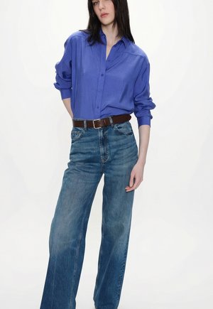 Frau trägt ein lockeres blaues Hemd mit Knöpfen, das in hoch taillierte, weite Jeans mit braunem Gürtel gesteckt ist, und steht vor einem weißen Hintergrund.